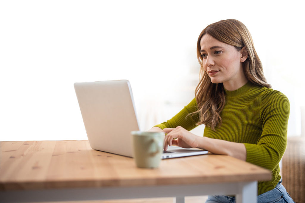 A white woman in a green top sits at a dining table with a laptop and mug, reading something from her screen.