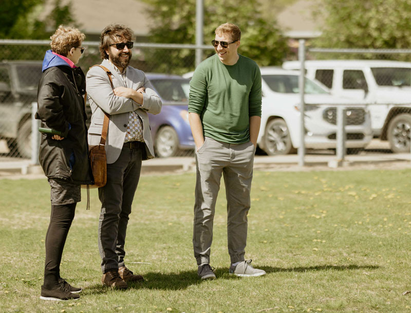 Two men and a woman talk while standing in a school yard and observing a student activity.