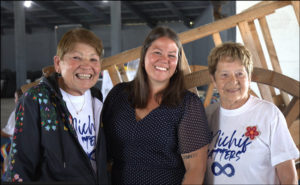 Three women smile while standing in front of a Red River cart.