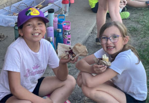 Two children sit on the ground eating bannock with jam.