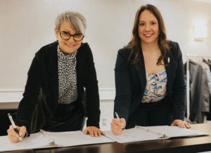 Two women stand at a table to sign a document.