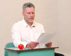 A male teacher sits in a school desk reading a letter.