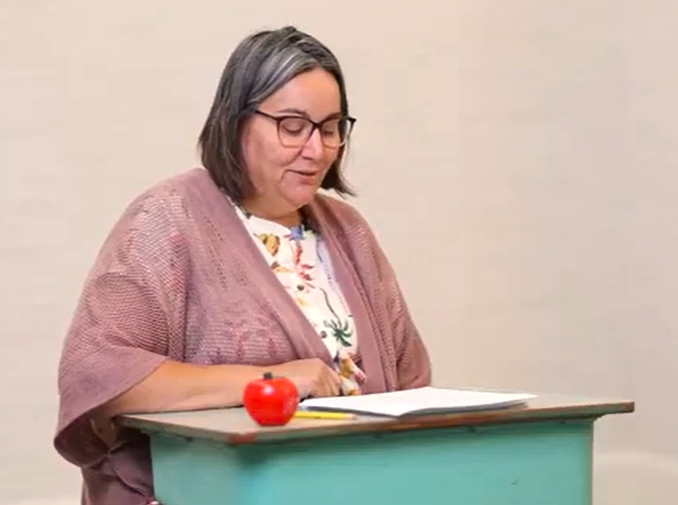 A female teacher sits in a school desk reading a letter.