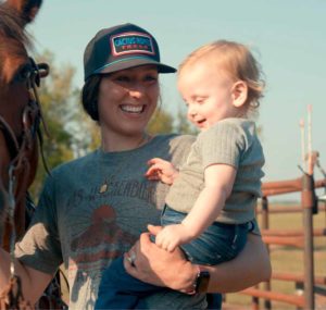 A woman holds a young child in one arm and the reins of a horse with her other hand.