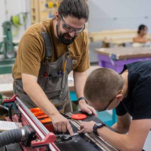 A student uses a wrench to tighten a bit on a router table while a teacher looks on.