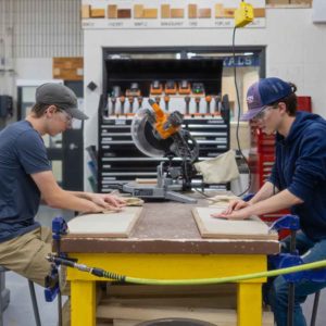Two male teenagers sand a wood project while sitting at a table.