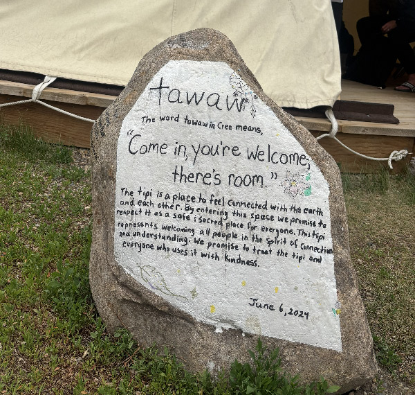 A painted rock outside a tipi. The hand lettered inscription reads: tawaw - The word tawaw in Cree means, 'Come in, you're welcome; there's room.' The tipi is a place to feel connected with the earth and each other. By entering this space we promise to respect it as a safe and sacred place for everyone. This tipi represents welcoming all people in the spirit of connection and understanding. We promise to treat the tipi and everyone who uses it with kindness. - June 6, 2024